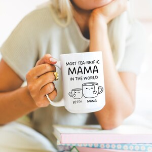 Może przedstawiać: Biały kubek ceramiczny z czarnym tekstem, kt&oacute;ry brzmi "Most tea-rrific Mama in the world." Kubek przedstawia dwie kresk&oacute;wkowe filiżanki do herbaty z twarzami, jedna oznaczona jako "Betty", a druga jako "Mama."