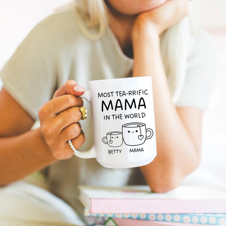 Może przedstawiać: Biały kubek ceramiczny z czarnym tekstem, kt&oacute;ry brzmi "Most tea-rrific Mama in the world." Kubek przedstawia dwie kresk&oacute;wkowe filiżanki do herbaty z twarzami, jedna oznaczona jako "Betty", a druga jako "Mama."