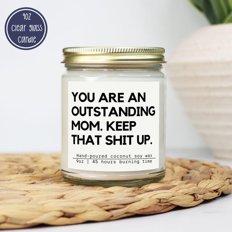 May include: A clear glass candle jar with a gold lid. The label reads "YOU ARE AN OUTSTANDING MOM. KEEP THAT SHIT UP." The candle is hand-poured coconut soy wax, 255g, and has a 45-hour burn time. A blue circle with text is in the upper left.