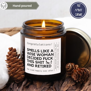 May include: A lit amber glass candle with a white label that reads "Smells Like a Wise Woman Decided Fuck This Shit and Retired." The candle is 4oz and hand-poured with coconut soy wax. The candle is surrounded by pinecones.
