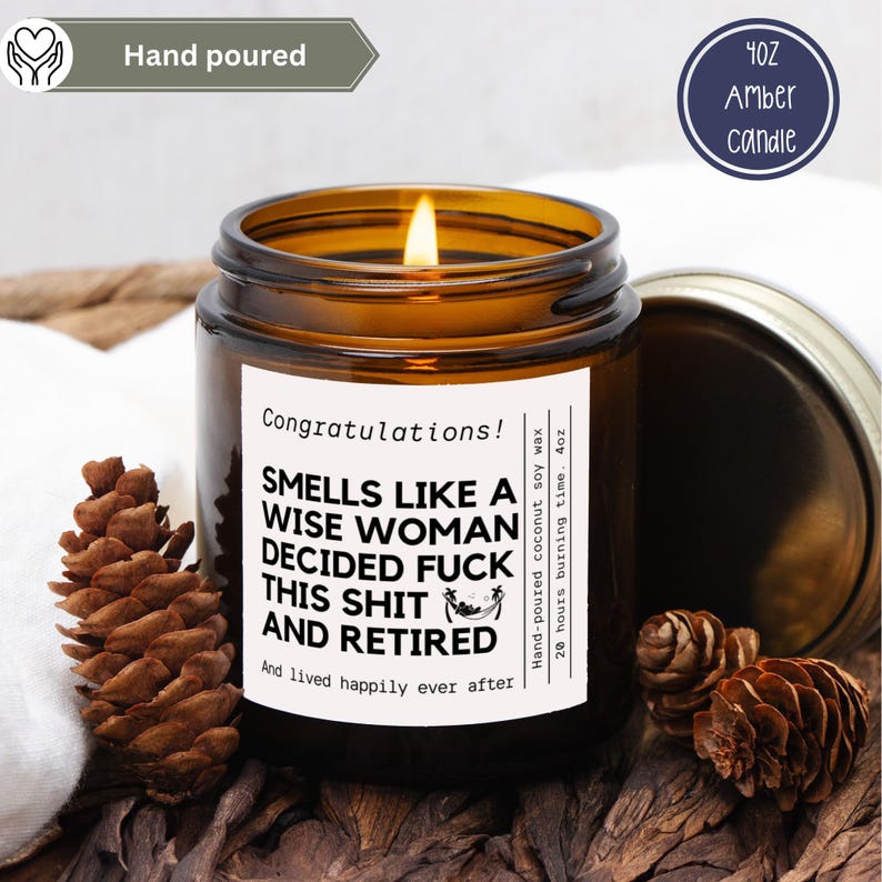 May include: A lit amber glass candle with a white label that reads "Smells Like a Wise Woman Decided Fuck This Shit and Retired." The candle is 4oz and hand-poured with coconut soy wax. The candle is surrounded by pinecones.