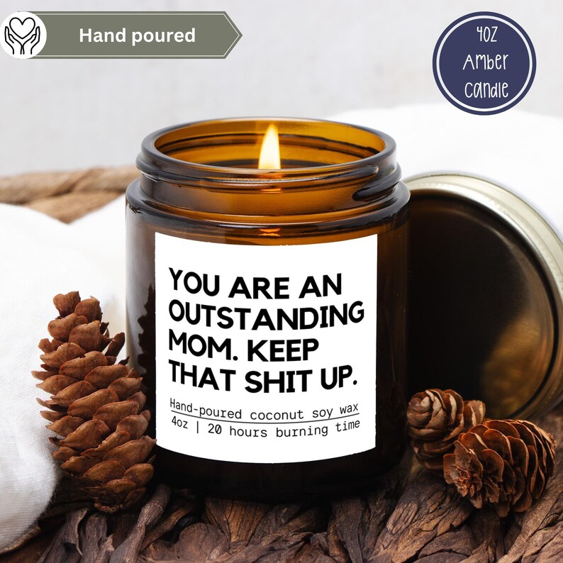 May include: An amber glass candle with a lit wick, labelled "YOU ARE AN OUTSTANDING MOM. KEEP THAT SHIT UP." The candle is hand-poured, made with coconut soy wax, and has a 20-hour burn time. It is 113 grams. Pine cones are nearby.