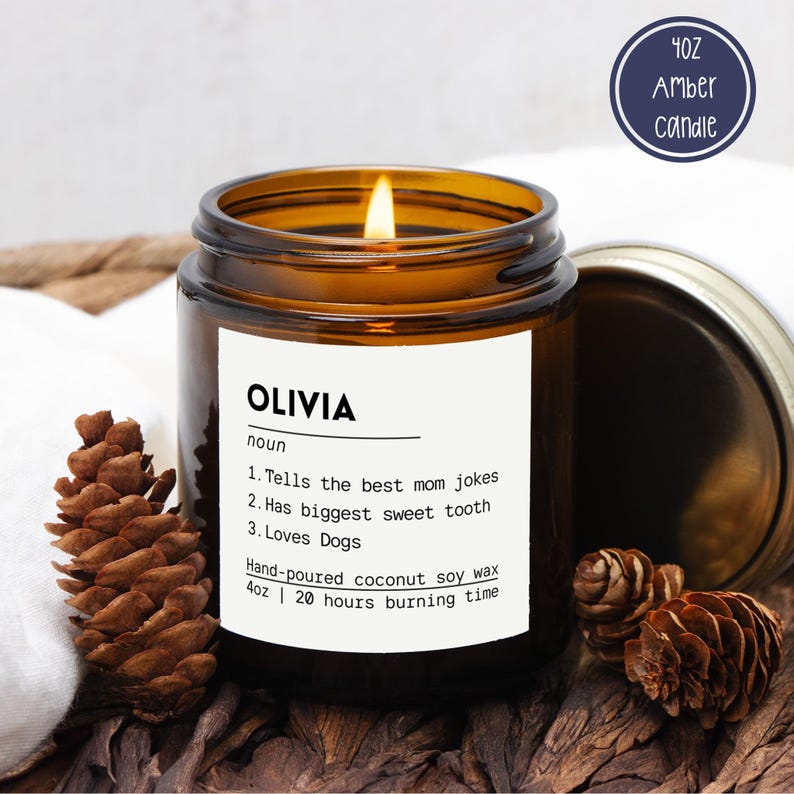 May include: A 4oz amber glass candle with a lit wick and a white label reading OLIVIA noun 1. Tells the best mom jokes 2. Has biggest sweet tooth 3. Loves Dogs. Hand-poured coconut soy wax, 4oz, 20 hours burning time. Includes a metal lid.