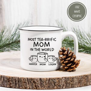 Może przedstawiać: Biały kubek ceramiczny z czarną obw&oacute;dką i uchwytem. Kubek ma czarny wz&oacute;r z napisem "Most Tea-Rrific Mom in the World" z trzema kresk&oacute;wkowymi filiżankami do herbaty pod spodem. Filiżanki do herbaty są oznaczone jako "Harper", "Mom" i "Logan".