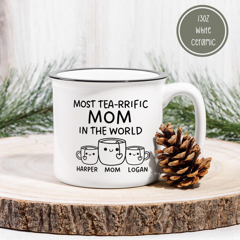Może przedstawiać: Biały kubek ceramiczny z czarną obw&oacute;dką i uchwytem. Kubek ma czarny wz&oacute;r z napisem "Most Tea-Rrific Mom in the World" z trzema kresk&oacute;wkowymi filiżankami do herbaty pod spodem. Filiżanki do herbaty są oznaczone jako "Harper", "Mom" i "Logan".