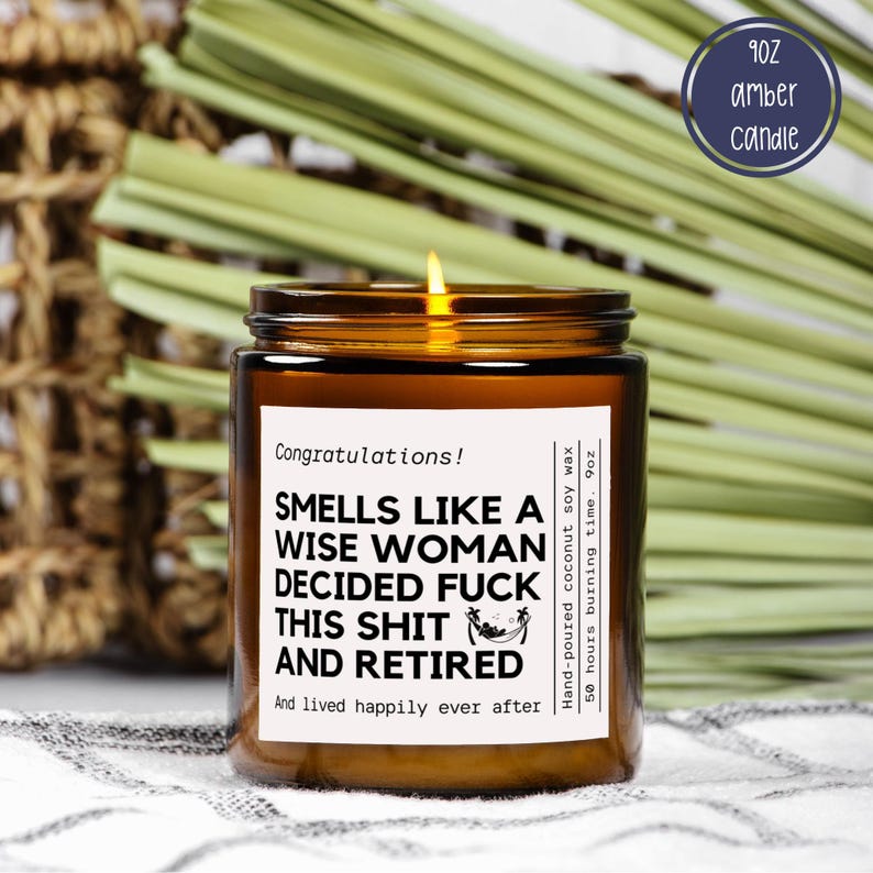May include: A lit amber glass candle with a white label that reads "Smells like a wise woman decided fuck this shit and retired." The candle is 9 oz and made with hand-poured coconut soy wax. The candle has a 50-hour burn time.