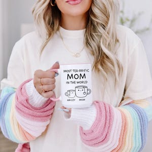 Może przedstawiać: Biały kubek ceramiczny z r&oacute;żowym rantem i uchwytem. Kubek ma czarny wz&oacute;r z napisem "Most Tea-rrific Mom in the World" z dwoma kresk&oacute;wkowymi filiżankami do herbaty, jedna z etykietą "Hailey", a druga "Mom".