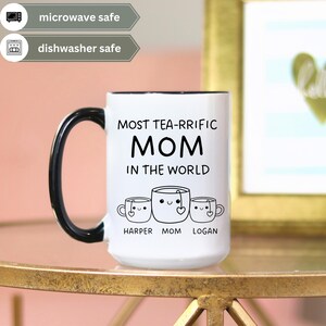 Może przedstawiać: Biały kubek ceramiczny z czarną obw&oacute;dką i uchwytem. Kubek ma czarny wz&oacute;r z napisem "Most Tea-rrific Mom in the World" z trzema kresk&oacute;wkowymi filiżankami do herbaty pod spodem. Filiżanki do herbaty mają napisane pod spodem imiona "Harper", "Mom" i "Logan".