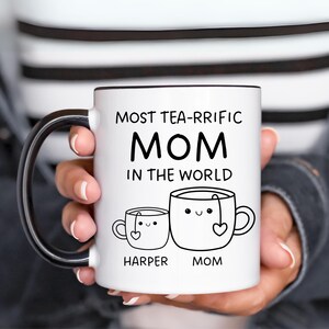 Może przedstawiać: Biały kubek ceramiczny z czarnym uchwytem. Kubek ma czarny napis "Most Tea-rrific Mom in the World". Pod napisem znajdują się dwie kresk&oacute;wkowe filiżanki do herbaty z twarzami i sercami. Mniejsza filiżanka ma napis "Harper", a większa "Mom".