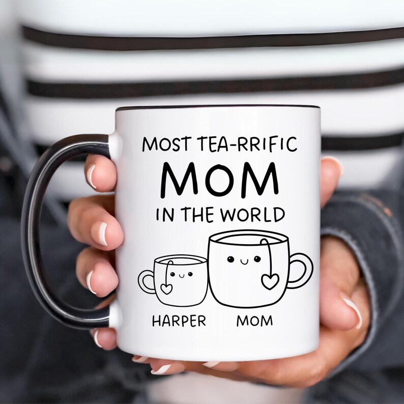 Może przedstawiać: Biały kubek ceramiczny z czarnym uchwytem. Kubek ma czarny napis "Most Tea-rrific Mom in the World". Pod napisem znajdują się dwie kresk&oacute;wkowe filiżanki do herbaty z twarzami i sercami. Mniejsza filiżanka ma napis "Harper", a większa "Mom".