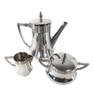 Puede incluir: Un juego de té plateado que incluye una cafetera, una lechera y un azucarero. La cafetera tiene un diseño alto y esbelto con tapa y un pico largo. La lechera y el azucarero tienen detalles a juego. El juego es de metal brillante.
