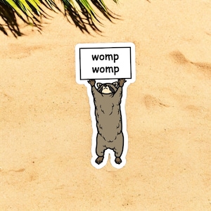 May include: A cartoon raccoon holding a sign that says "womp womp" on a sandy beach background.