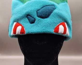 May include: A green and blue fleece hat with a cartoon character design. The character has red eyes and a white mouth.