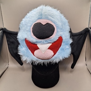 May include: A whimsical, plush hat in the shape of a cartoon monster. The hat features a light blue, furry exterior with a large, black eye, a red smiling mouth, and black bat wings. The hat is designed to be worn on the head.