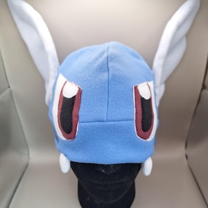 Pode incluir: Um gorro de tecido polar azul-claro com duas orelhas brancas grandes e verticais nas laterais. A frente do gorro exibe dois olhos grandes com seções brancas, pretas e vermelho-escuras, além de duas pequenas presas brancas na borda.