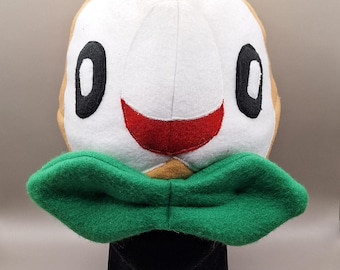 May include: A plush toy shaped like a round bird head with tan and white fabric. It features large black oval eyes, a small red mouth, and a prominent green fabric bow tie attached at the base of the face. The design is soft and rounded.