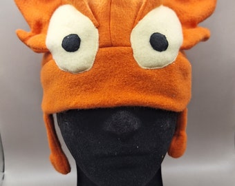 May include: Orange monster hat with large white and black eyes, and ear flaps. The hat is made of a soft, felt-like material. The hat is displayed on a black mannequin head.