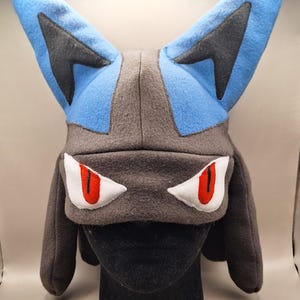 Puede incluir: Un gorro de forro polar diseñado para parecerse a una criatura con grandes orejas puntiagudas azules y grises. El gorro presenta una base gris oscuro con aplicaciones en forma de ojos blancos y rojos en el frente y solapas laterales largas.