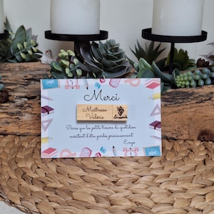 Peut inclure: Une clé USB en bois avec les mots "Merci" et "Maîtresse Valérie" gravés sur une carte à fond bleu clair. La carte a une bordure décorative avec des illustrations sur le thème de l'école. Le texte "Parce que les petits trésors du quotidien méritent d'être gardés précieusement Enzo" est également visible.