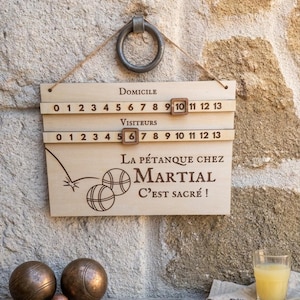Peut inclure: Panneau en bois avec anneau métallique pour accrocher, avec des compteurs de points pour domicile et visiteurs. Le panneau indique "La Pétanque chez Martial, C'est Sacré !" avec des illustrations de boules de pétanque. Boules de pétanque en bronze.