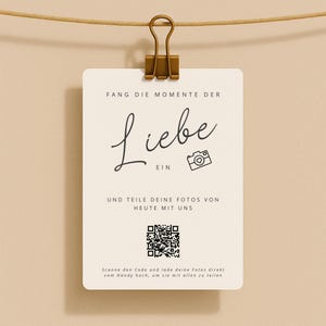 Könnte beinhalten: Eine cremefarbene Karte mit dem Wort "Liebe" in eleganter Schrift, gehalten von einer goldenen Klammer. Die Karte enthält deutschen Text, ein Kamera-Symbol und einen QR-Code. Die Karte hängt an einer braunen Schnur vor beigem Hintergrund.