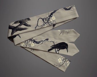 May include: A beige fabric sash with a black crow and white flower design. The sash is folded in thirds and has a subtle sparkle.