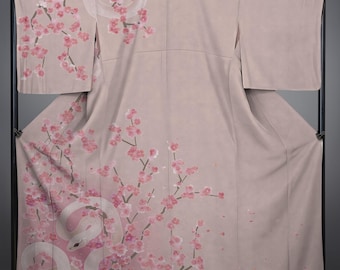 May include: A light pink kimono with a white snake design intertwined with pink cherry blossom branches. The kimono is displayed on a black metal rack, showcasing its flowing fabric and intricate artwork. The design is repeated on the sleeves.