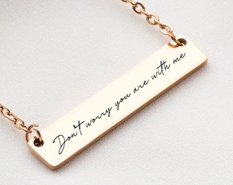 May include: A gold-colored bar necklace with a delicate chain. The rectangular pendant features the inscription "Don't worry you are with me" in elegant script. The necklace is displayed on a white surface.
