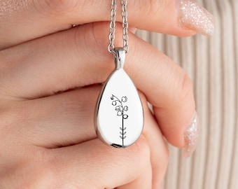 May include: Silver-toned teardrop pendant necklace featuring a delicate floral design. The pendant hangs from a fine chain. The floral design is a black line drawing of a flower with small bell-shaped blooms.