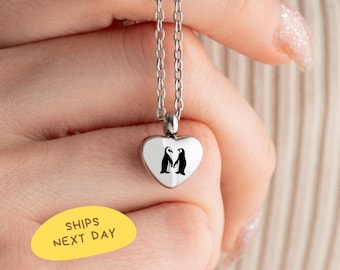 May include: Silver-toned necklace with a heart-shaped pendant. The pendant features a black penguin design. The necklace is displayed against a neutral background. The text "SHIPS NEXT DAY" is on a yellow oval.