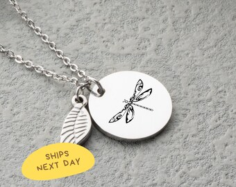 May include: A silver-colored necklace with a dragonfly pendant. The pendant is a white, circular disc with a black dragonfly design. A small leaf charm and a yellow oval with the text "SHIPS NEXT DAY" are also attached.