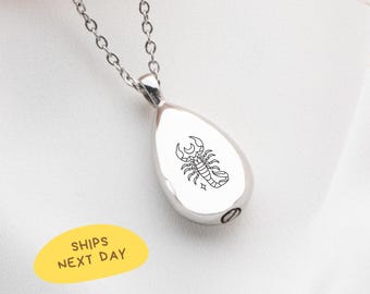 May include: A silver-colored teardrop pendant necklace with a black scorpion zodiac symbol. The pendant hangs from a delicate chain. The pendant is approximately 1 inch in length. The background is a white fabric.
