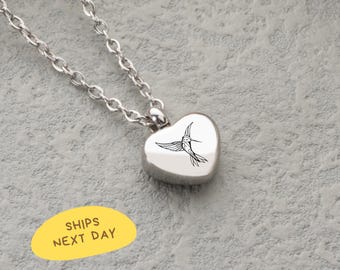 May include: A silver-colored heart-shaped pendant necklace with a hummingbird design. The pendant hangs from a delicate chain. The hummingbird is depicted in black ink. The necklace is displayed on a textured gray surface.