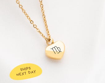 May include: A gold-colored necklace with a heart-shaped pendant. The pendant features the Virgo symbol in black. The necklace chain is also gold. The image includes a yellow oval with the text "SHIPS NEXT DAY".
