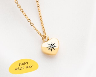 May include: A gold-colored heart-shaped pendant necklace with a black sunburst design. The necklace has a delicate chain and is displayed on a white background. The text "SHIPS NEXT DAY" is on a yellow oval.