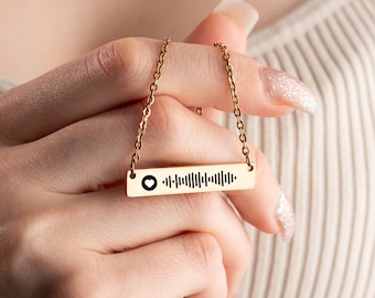 May include: Gold-colored necklace with a rectangular pendant featuring a black heart and a series of vertical lines, resembling a barcode. The necklace is held by a person, showcasing the delicate chain and pendant design. The background is a neutral beige.