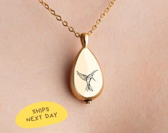 May include: Gold-colored teardrop pendant necklace with a hummingbird design. The pendant hangs from a delicate chain. The hummingbird is black and white. The necklace is worn on a person's neck.