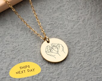 May include: Gold-colored necklace with a round pendant featuring a line drawing of hands cradling baby feet. The chain is delicate, and the pendant is centered. The background is a textured gray surface.