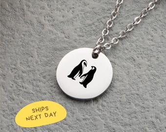May include: A silver-colored necklace featuring a circular pendant with a black penguin design. The pendant is attached to a delicate chain. The background is a textured gray surface.