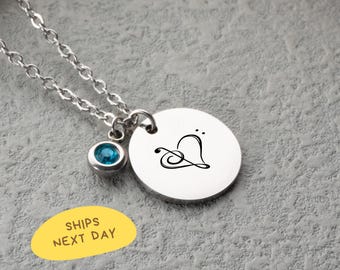 May include: A silver necklace with a round pendant featuring a black heart design. A small, round, blue gemstone hangs below the chain. The necklace is on a gray surface. The text "SHIPS NEXT DAY" is on a yellow oval.