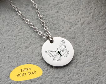 May include: A silver-colored necklace with a circular pendant featuring a black butterfly design. The chain is made of small, linked metal pieces. The pendant is approximately 1 inch in diameter. The text "SHIPS NEXT DAY" is on a yellow oval.