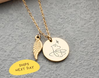 May include: Gold-colored necklace with a chain and two charms. One charm is a wing, and the other is a round pendant with a cat illustration. The text "SHIPS NEXT DAY" is on a yellow oval.