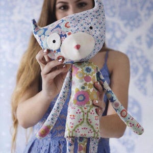 May include: A fabric doll featuring a cat-like head with pointed ears, a white muzzle, and a small pink nose. The doll is constructed from various floral patterned fabrics in shades of blue, pink, green, and white, with long slender arms and legs.
