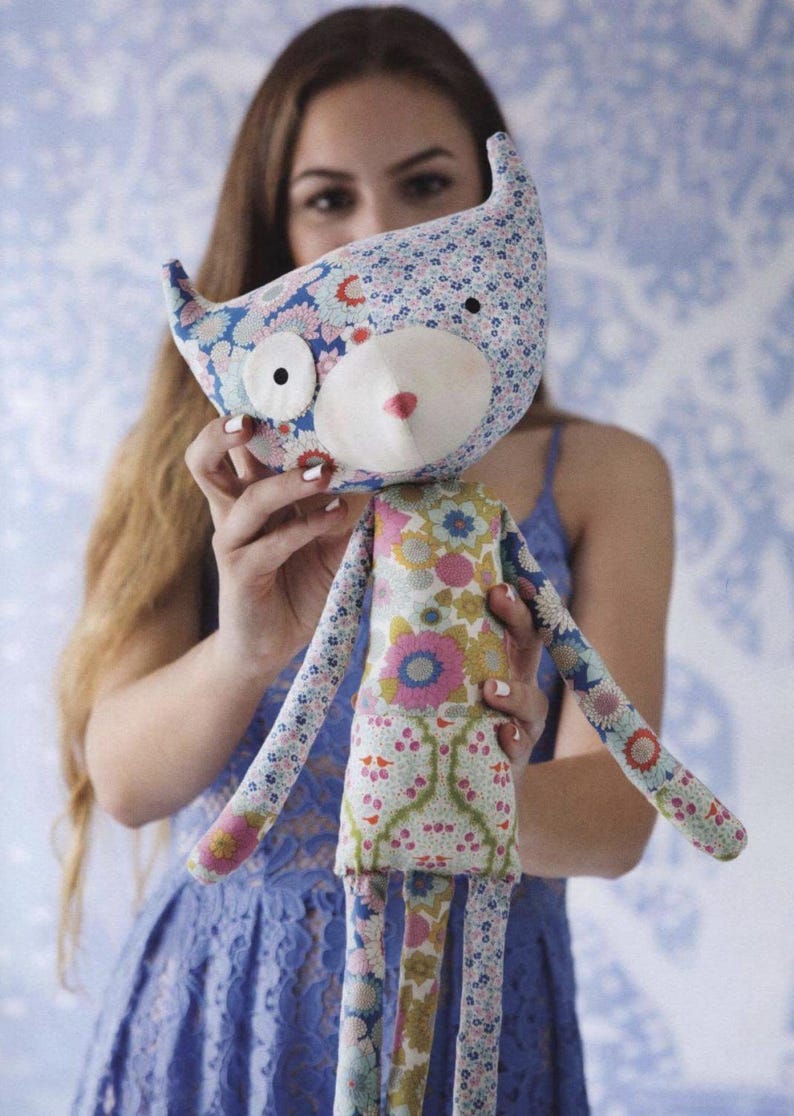 May include: A fabric doll featuring a cat-like head with pointed ears, a white muzzle, and a small pink nose. The doll is constructed from various floral patterned fabrics in shades of blue, pink, green, and white, with long slender arms and legs.