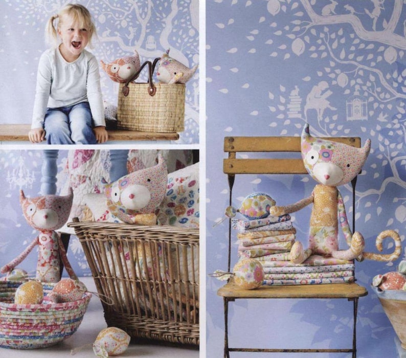 May include: A collection of handcrafted fabric cat dolls featuring various floral and patterned textiles in soft pastel colors. The dolls have pointed ears, long tails, and stitched facial features, displayed alongside woven baskets and a wooden chair.