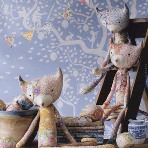 May include: Three handmade fabric fox dolls crafted from various floral and patterned cotton patches. Each fox features pointed ears, a solid cream-colored snout, and small black eyes. The dolls are positioned around a wooden bowl and stack of fabric.