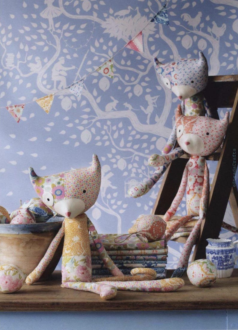 May include: Three handmade fabric fox dolls crafted from various floral and patterned cotton patches. Each fox features pointed ears, a solid cream-colored snout, and small black eyes. The dolls are positioned around a wooden bowl and stack of fabric.