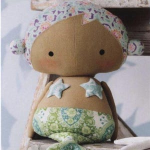 May include: A plush mermaid doll with tan skin, two small black eyes, and pink cheeks. The doll features a floral patterned head covering, two light blue star-shaped patches on the chest, and a green and white paisley patterned tail fin.