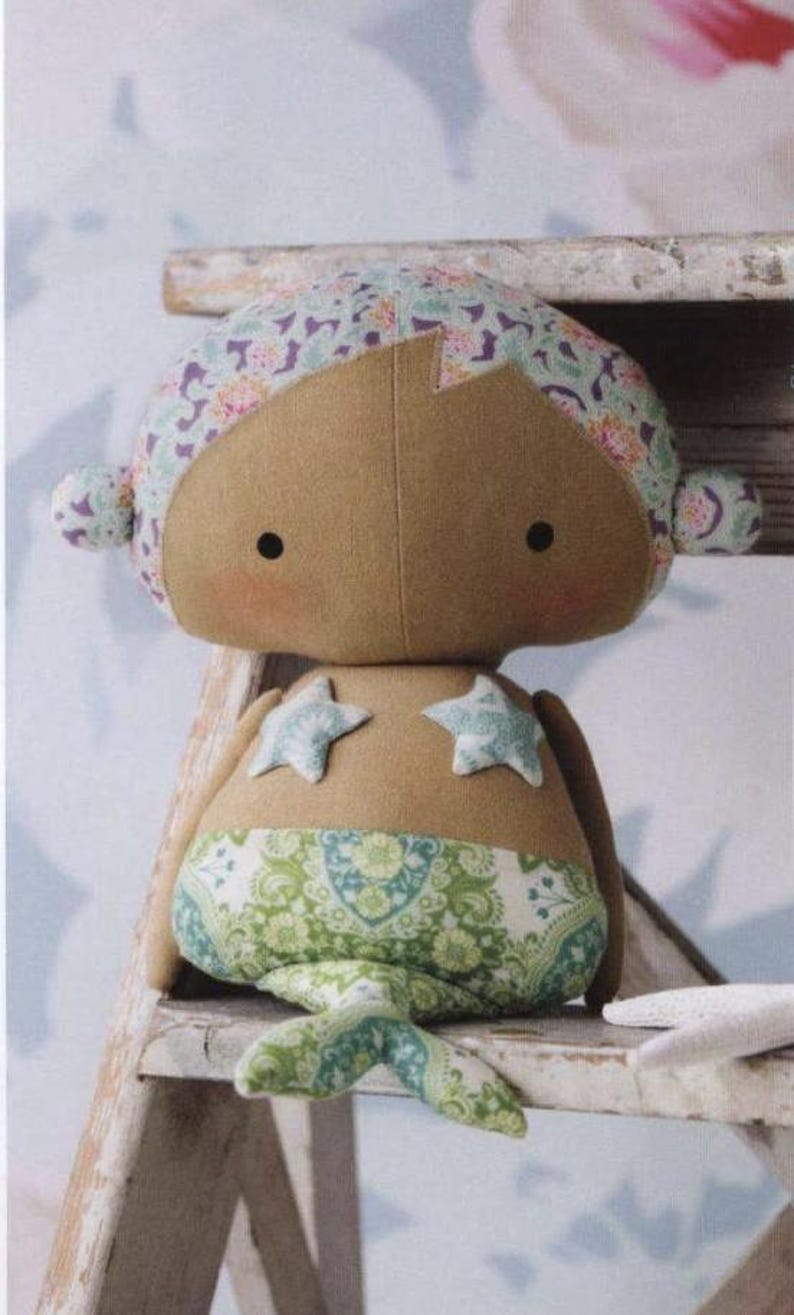 May include: A plush mermaid doll with tan skin, two small black eyes, and pink cheeks. The doll features a floral patterned head covering, two light blue star-shaped patches on the chest, and a green and white paisley patterned tail fin.