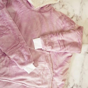Puede incluir: Una sudadera con capucha de gran tama&ntilde;o hecha de felpa suave en color malva. La prenda cuenta con un forro grueso de sherpa color crema dentro de la capucha y un bolsillo canguro frontal. Tiene mangas largas con pu&ntilde;os blancos acanalados.
